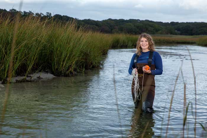 Faith Brooks, wearing waders, stands knee-deep in a pond, holding an instrument and a coil of rope.